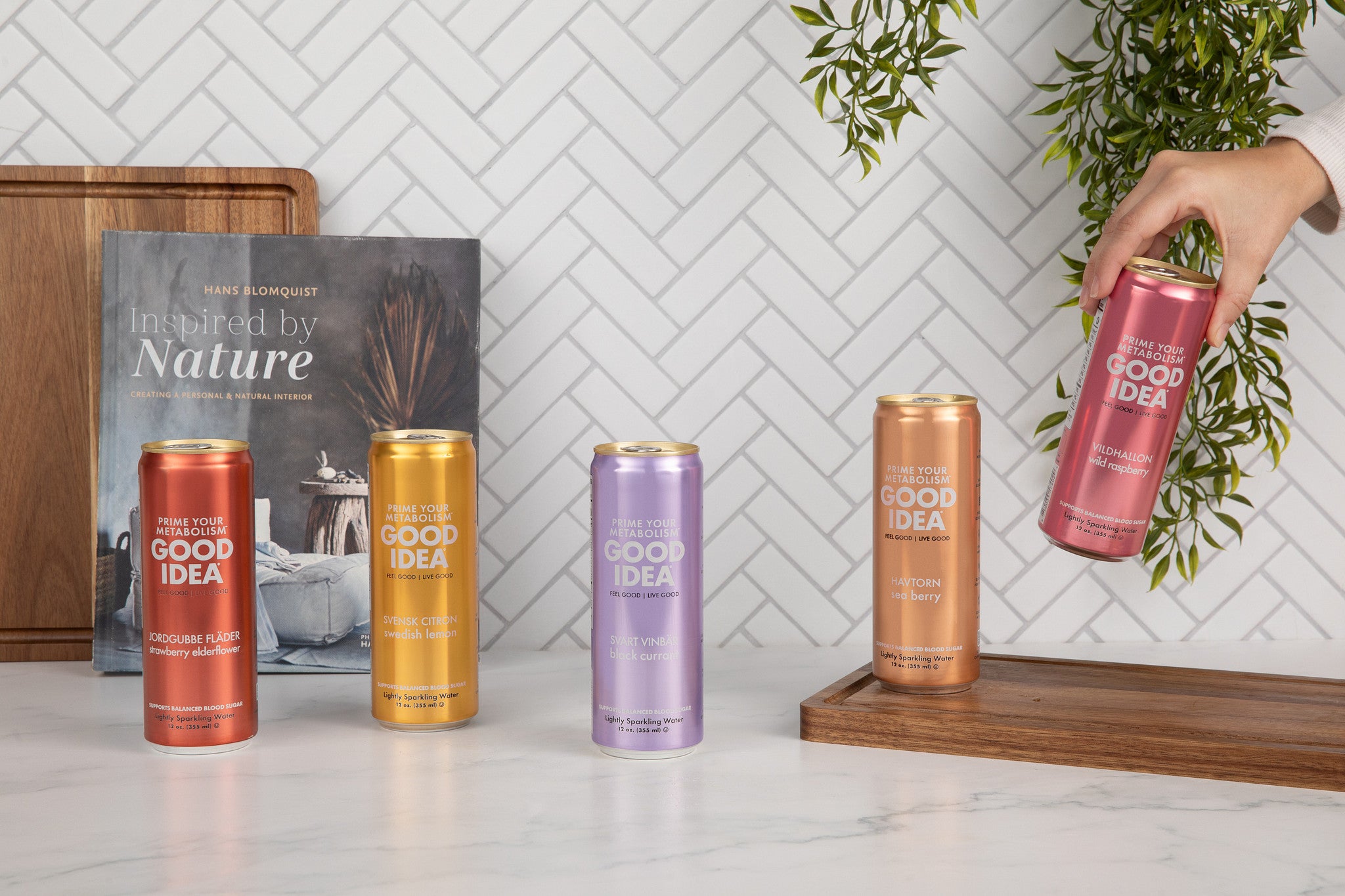 All five Good Idea cans on a countertop with a person picking up the Wild Raspberry flavor