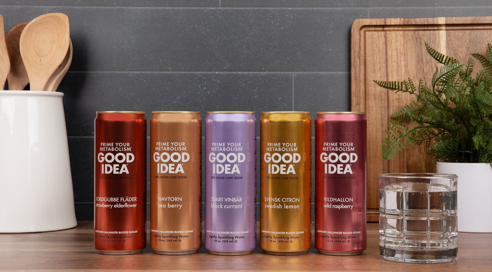 Five cans of Good Idea sparkling water on a kitchen counter, with a plant and cutting board in the background. A glass of sparkling water sits to the side.