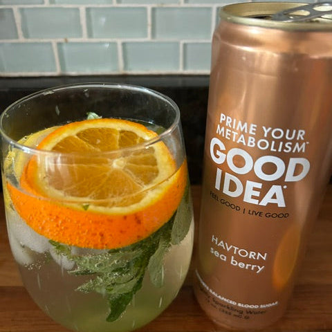 A can of GOOD IDEA Sea Berry drink is shown next to a glass of the beverage garnished with an orange slice and mint on a wooden surface.