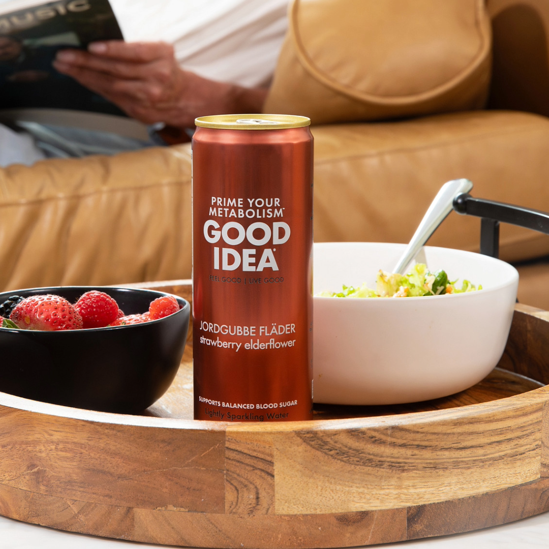 Can of GOOD IDEA on a wooden tray beside bowls of fresh fruit and salad, with a person reading in the background.